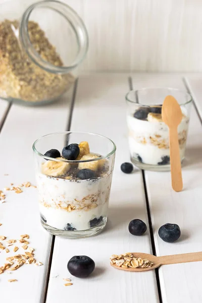 Dos vasos con yogur en capas, avena y frutas, sobre una mesa de madera blanca. Desayuno saludable, concepto de estilo de vida saludable —  Fotos de Stock