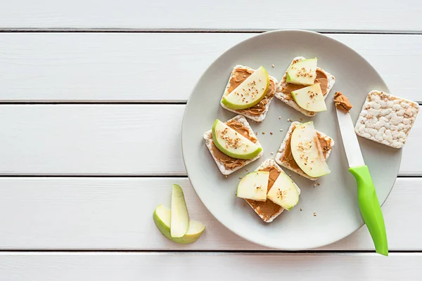 Healthy breakfast with peanut butter and apple sandwiches on rice cakes. Top view on white wooden background, copy space. — Stock Photo, Image