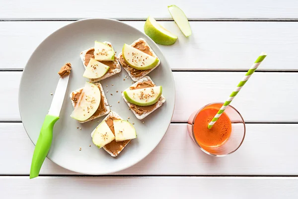Healthy breakfast with peanut butter and apple sandwiches on rice cakes, carrot juice. Top view on white wooden background. — Stock Photo, Image