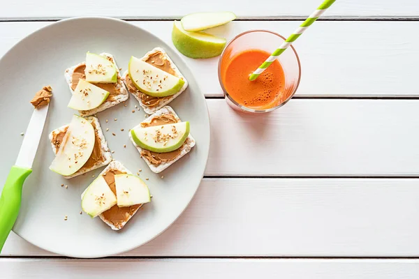 Healthy breakfast with peanut butter and apple sandwiches on rice cakes, carrot juice. Top view on white wooden background, copy space. — Stock Photo, Image