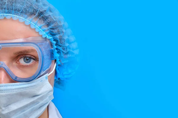 Sad doctor in a white medical coat and safety glasses, copy space on blue background. Concept stay home because of flu epidemic. Sad nurse in medical mask with a stethoscope.
