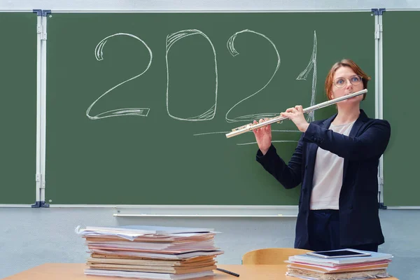 A music teacher plays a musical instrument on the background of a blackboard with the text 2021. . Learning to play wind instruments in quarantine during the coronavirus pandemic