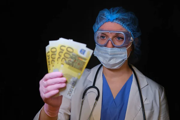 A woman medical doctor holds cash euros in her hands. A nurse girl with money in a protective mask on a black background. Concept of financial assistance to people under coronavirus quarantine