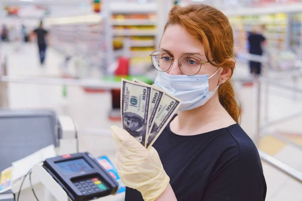 A female cashier in a medical mask counts money against the background of the cash register. Dollars in the hands of the seller in the store