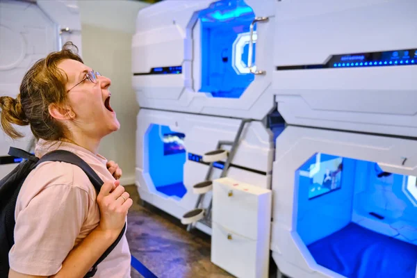A young woman yawns and wants to sleep next to the rooms of a mini hotel. Capsule hotel - one of the variants of high tech hotels, which is a small sleeping cells located on top of each other
