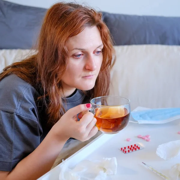 A woman with red eyes who is ill with the flu virus drinks tea