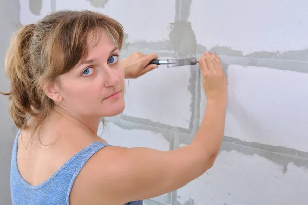 A young woman with a hammer looking at the camera. Hammering a nail into the wall of foam blocks in the new apartment. Repair yourself in the built house.