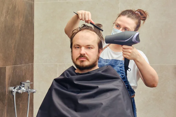 Woman with hairdryer doing hairstyle to man in home bathroom. Concept of recovery and return to normal life after the coronavirus epidemic