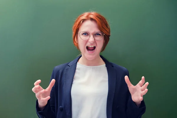 Woman teacher screams furious at the blackboard, copy space background. Angry school teacher on green background, close up