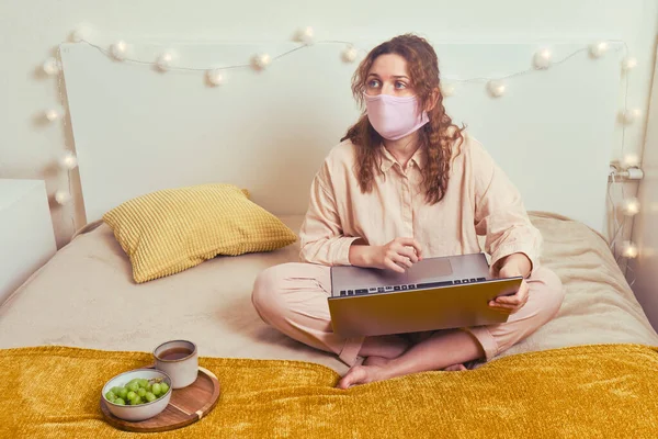 A woman in a medical mask works online with a laptop on a home bed. Remote work from home during lockdown due to coronavirus.