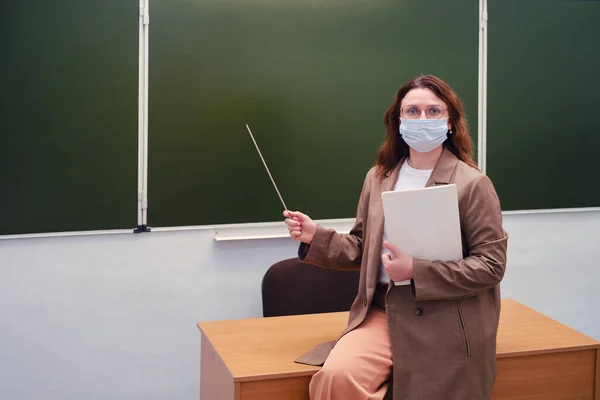 A teacher in a medical mask sits on a desk at the blackboard