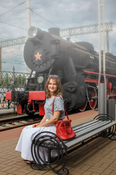 Young woman sitting on the platform near retro train. Girl with red bag on the bench near the steam locomotive. Cloudy weather and smoke from train at open station. Vintage style.