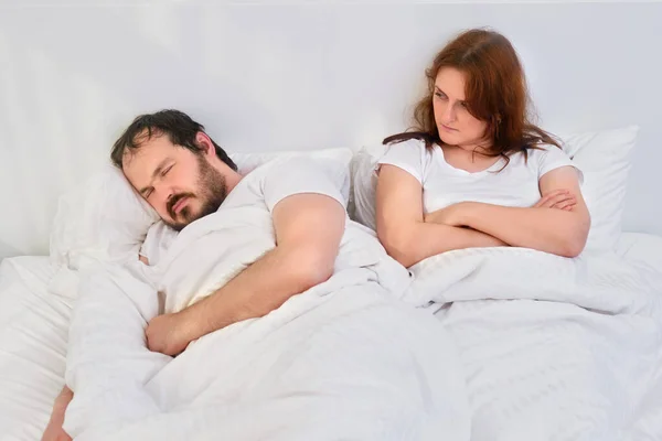 Angry woman looking at a man sleeping in a white bed, concept. Relationship problems coupled with isolation due to coronavirus.