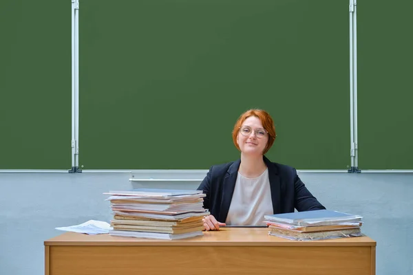 Woman teacher smiling sitting at her desk at the school blackboard. Happy school teacher, copy space on green background