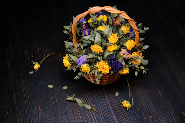 Bouquet of dried flowers on a wooden background. Dry yellow roses in a wicker basket. Beautiful faded flowers on the boards of black wood. Blue and yellow buds in vintage style.