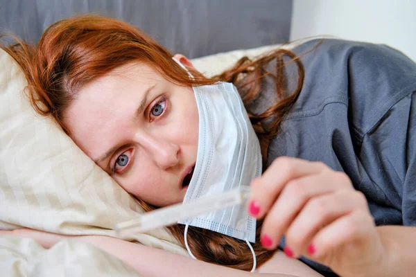 Redhead woman sick with a coronavirus in shock looks at a thermometer. Woman lies on a bed in a medical mask