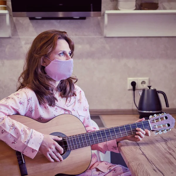 A young woman in a medical protective mask plays a home kitchen guitar. Classes over the Internet during quarantine due to coronavirus