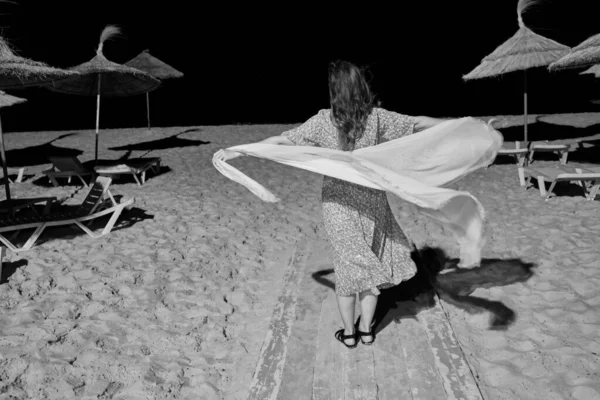 A woman with a flowing dress stands on a night beach, monochrome