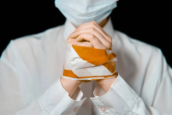 Hands of a man in a medical mask, bound with a stop-tape, close-up. Tied woman in a protective mask on a black background, the concept of a coronavirus quarantine
