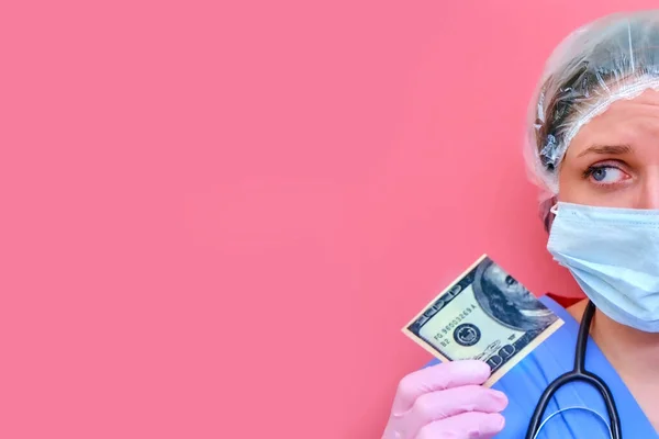 A woman medic holds a us dollar bill cut in half, copy space for text. A nurse with money for work on a pink background, the concept of paying doctors in a crisis due to the coronavirus epidemic