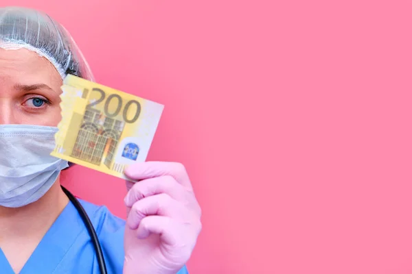 A female medic holds a Euro bill torn in half, copy space for text. A nurse with money for work on a pink background, the concept of paying doctors in a crisis due to the coronavirus epidemic.