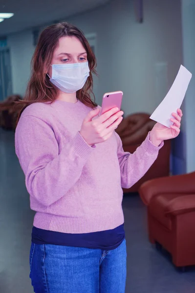 A woman in a medical mask with a phone in her hand was thinking in the hospital corridor. Patient holds a piece of paper with a doctor referral and watch online from their phone