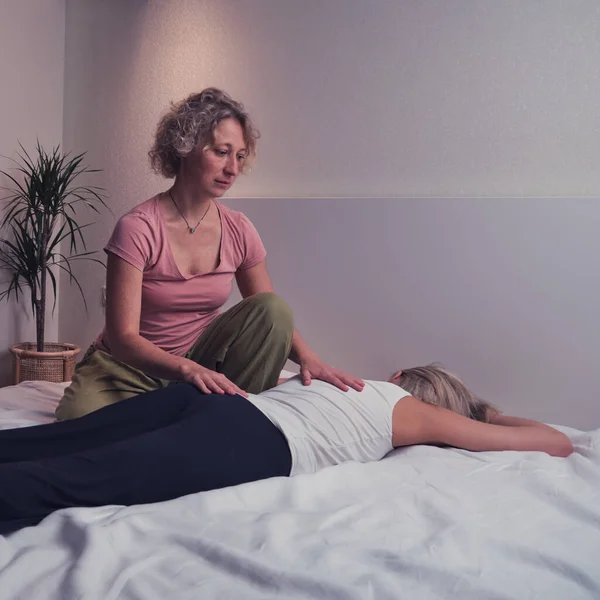 A woman does a holistic back massage on a home bed in the bedroom. Blonde masseuse on the couch massages a female client