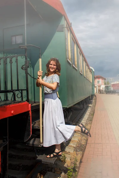 Woman standing on the running Board of the car. Holding the handrail of the departing train. Empty platform in vintage style. Retro cars and locomotive.