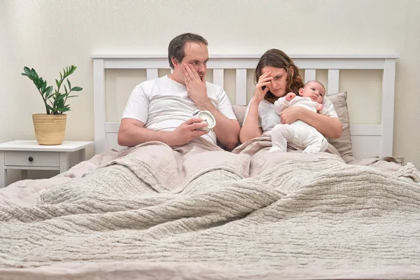 Sleepy mother and father with a baby boy, problems of parents with an infant child on a home bed. Difficulties of a man and a woman with a newborn baby in the bedroom