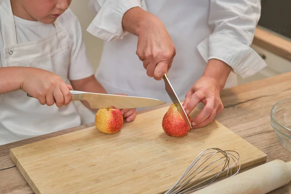 Mom and son in white chef\'s clothes cut pears with a knife in their home kitchen. An adult woman with a child boy will cook a pie