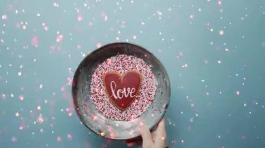 Romantik bir animasyon kutlama videosu. Tek bir kırmızı kalp, yumuşak mavi arka planda beyaz bir kasenin içinde duruyor. Birdenbire renkli konfeti parçacıkları, Sevgililer Günü için mükemmel bir şenlik sağanağında kalpten yukarıya doğru patlar.,