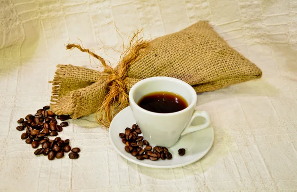 Bodegón: taza blanca de café negro con saco de arpillera de granos de café tostados en el mantel de lino blanco — Foto de Stock