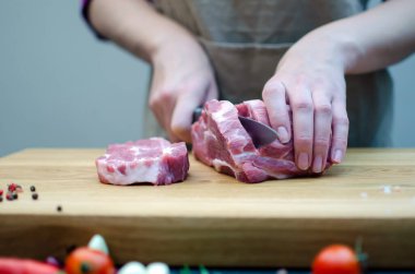 Mutfaktaki hostes domuz etini bıçakla keser, yemek hazırlar. Kesme tahtasında, baharatlar, biber, biberiye, sarımsak, kiraz domatesleri.