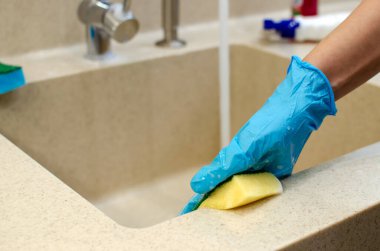 Kadın evdeki mutfakta ev işi yapıyor, lavabo ve musluğu sarı bir sünger, köpük ve suyla temizliyor. Katı yüzeyden yapılmış tezgah ve lavabo, mermer.