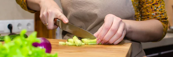 Vista ravvicinata del processo di preparazione dell'insalata. Una donna in grembiule taglia un cetriolo su un tagliere di legno — Foto Stock