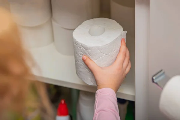 Rolo de papel higiênico branco na mão. A mão das crianças tira um rolo de papel higiénico do armário. Lotes de papel higiênico no fundo — Fotografia de Stock