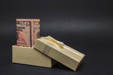 Hediye olarak para. Siyah homojen bir arka planda, fiyonklu bir kutuda bir dolar. Metnin için yer.