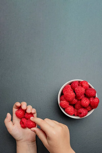 Raspberries in a saucer on a black background. The child takes raspberries from the plate. Shale stone texture. Vertical photo — Stock Photo, Image