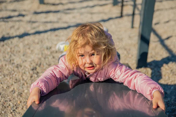 Uma menina brincando no playground sobe a colina. Uma atividade saudável de primavera ou outono para crianças. A criança joga alegremente ao ar livre. — Fotografia de Stock