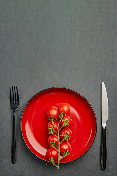 Помидоры в тарелке в качестве диеты или здорового питания концепции. Помидоры черри на красной тарелке с вилкой и ножом на черном каменном фоне. Место для текста или дизайна — стоковое фото