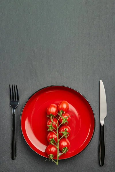 Tomaten auf dem Teller als Diät oder gesundes Ernährungskonzept. Kirschtomaten auf einem roten Teller mit Gabel und Messer auf schwarzem Steingrund. Platz für Text oder Design — Stockfoto