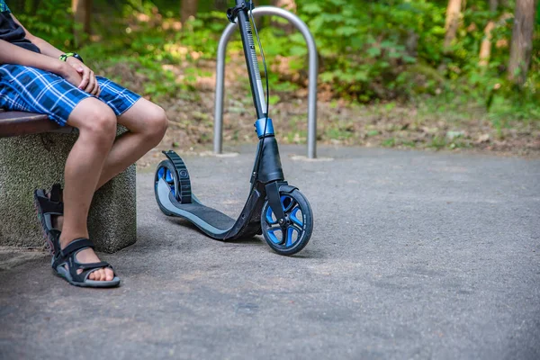 Gehen Sie im Park auf einem Motorroller. Ein junger Mann sitzt auf einer Bank und ruht sich aus, während er auf einem Smokat geht. Ökologie und ökologisches Verkehrskonzept — Stockfoto