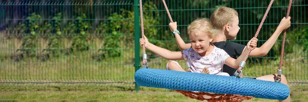 Gyerekek hintáznak. Fiú és lány hintáznak a parkban egy nyári napon. Hintázz a vidámparkban. banner szöveggel — Stock Fotó