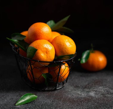 Yeşil yapraklı mandalinalar. Yapraklı parlak turuncu mandalinalar koyu bir arka planda sepette yatıyor. Karanlık fotoğraf. Fotoğraf desenli bir arkaplan gösteriyor.