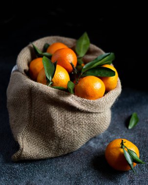 Yeşil yapraklı mandalinalar. Yapraklı parlak turuncu mandalinalar arka planda bir çuval içinde yatıyor. Noel meyvesi. Karanlık fotoğraf. Fotoğraf desenli bir arkaplan gösteriyor.