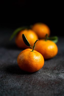 Yeşil yapraklı mandalinalar. Koyu dokulu yüzeyde yaprakları olan dört portakal rengi mandalina. Seçici odaklanma. Karanlık fotoğraf. Fotoğraf desenli bir arkaplan gösteriyor.