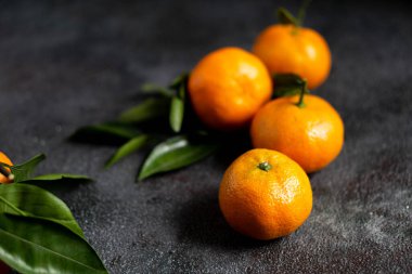 Yeşil yapraklı mandalinalar. Koyu dokulu yüzeyde yaprakları olan dört portakal rengi mandalina. Seçici odaklanma. Karanlık fotoğraf. Fotoğraf desenli bir arkaplan gösteriyor.