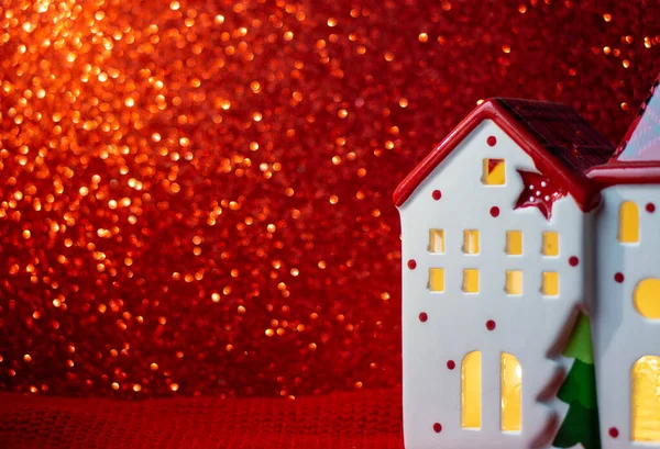 A festive figurine of a small house with glowing windows. Christmas decorations. New Year. Merry Christmas and new year, greeting card. Decorative house on a red background and gold bokeh. Copy space.