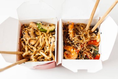 Karton kutuda kızarmış tavuk, deniz ürünü ve sebzeli Udon eriştesi ve beyaz arka planda ahşap yemek çubukları. Düz yatıyordu