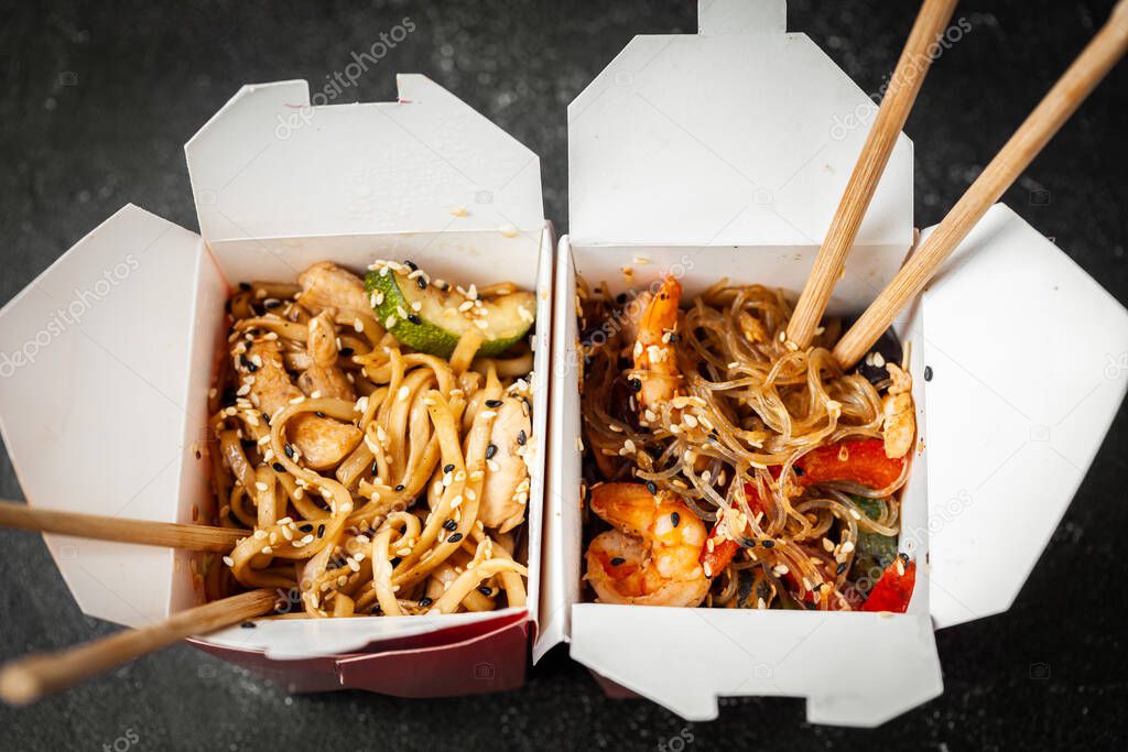 Udon noodles with fried chicken, seafood and vegetables in the cardboard box with wooden chopsticks on the black background. Flat lay